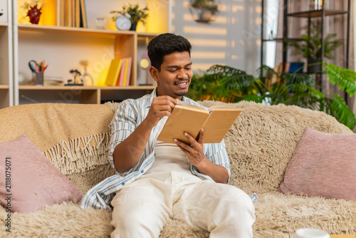 Indian man on home sofa reads a few lines in a book turns page and smiles at rest in living room. Arabian guy on home sofa enjoys quiet reading time and peaceful home leisure with warm relaxed mood.