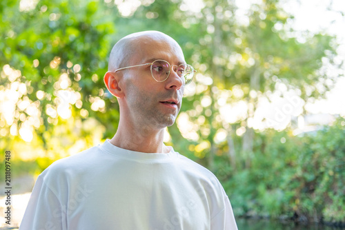 Young adult man with round glasses white t-shirt gesturing hand outdoors, discussing or debating topic, Modern male portrait against lush green foliage background, expression, casual streetwear style