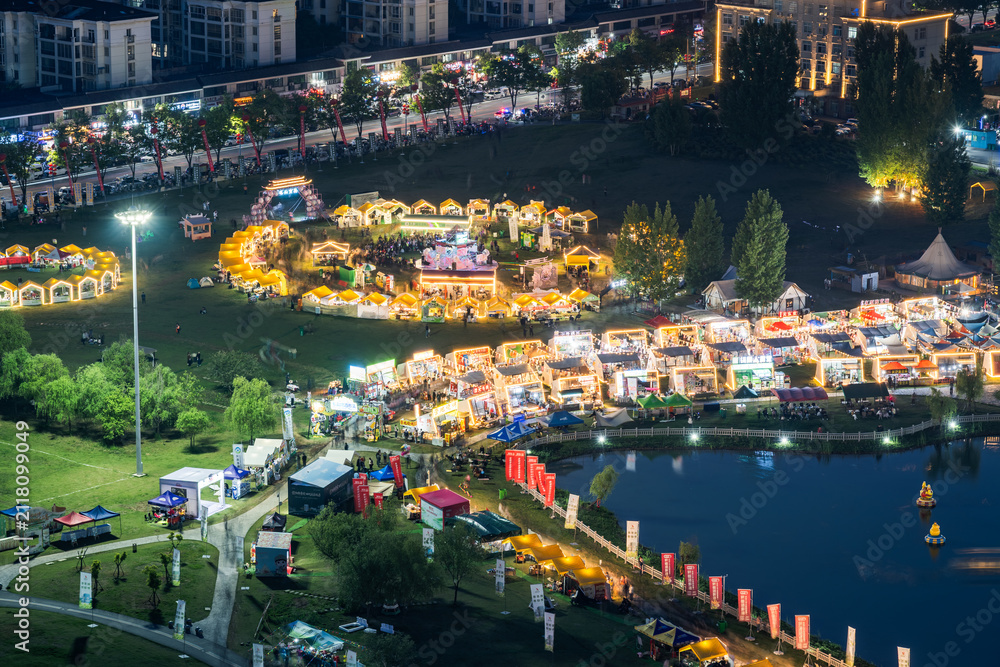 Fototapeta premium An illuminated outdoor night market and festival event set by a lake in an urban park, with stalls and event structures at night.