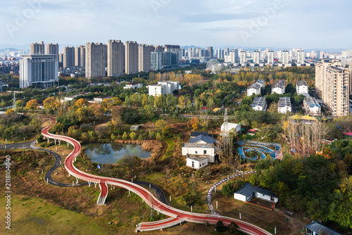 Photography Aerial view of an urban park with elevated red walkway, lake, trees, and surrounding residential high-rise buildings