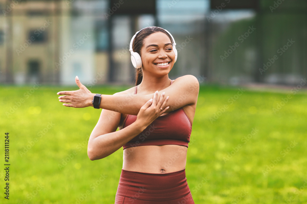 Fototapeta premium Smiling Asian Woman Stretching Arm While Listening To Music With Headphones In Park, Engaged In Outdoor Fitness Routine, Enjoying Active Lifestyle Surrounded By Nature. Weight Loss Concept.