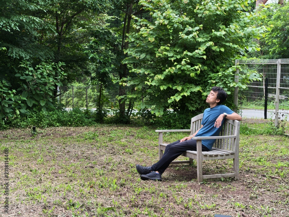 Fototapeta premium Man relaxes on wooden bench in peaceful garden. Greenery surrounds him, creating calm atmosphere. He leans back, eyes closed, appearing serene and at ease