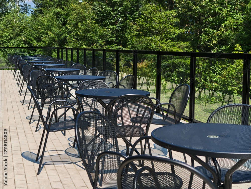 Fototapeta premium Rows of dark metal tables and chairs sit empty on a paved terrace. Green trees frame a black railing beyond the patio. The sun glints off the tables' smooth surfaces. No people occupy the space