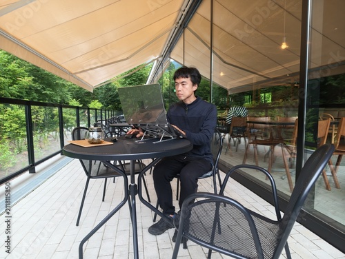 Young man uses laptop outdoors on terrace. Green trees frame glass walls behind him. Natural light highlights his focused expression. Table and chairs surround him on tiled floor
