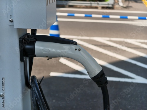 Electric vehicle charger plugged into a station. White and black cable rests against grey wall. Blue and white barriers mark parking zone. Sunlight casts clear shadows on asphalt