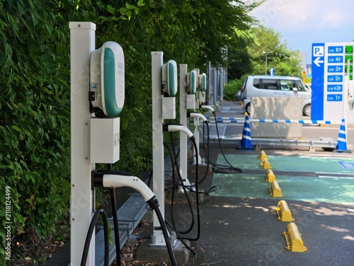 Row of modern electric vehicle chargers lines the park. Greenery borders the charging station neatly. Yellow curbs guide traffic along the paved path