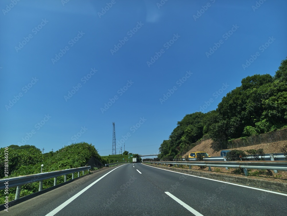 Fototapeta premium Clear blue sky stretches above winding asphalt road. Green trees line both sides, offering natural shade. Railing guards the edge, ensuring driver safety