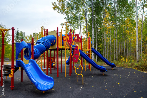 Colorful new playground with soft rubber pavement in the rural park in autumn, yellow and green trees around, no people.