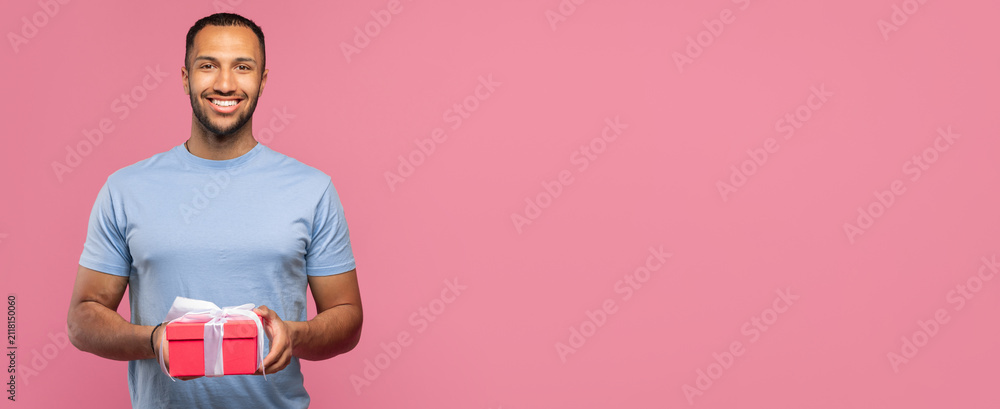 Obraz premium Smiling Arab Man In His 20s Holding A Gift Box With A Ribbon In Front Of Pink Background, Expressing Joyful Emotion. Engaged In Celebratory Activity In A Festive Setting. Celebration Concept.
