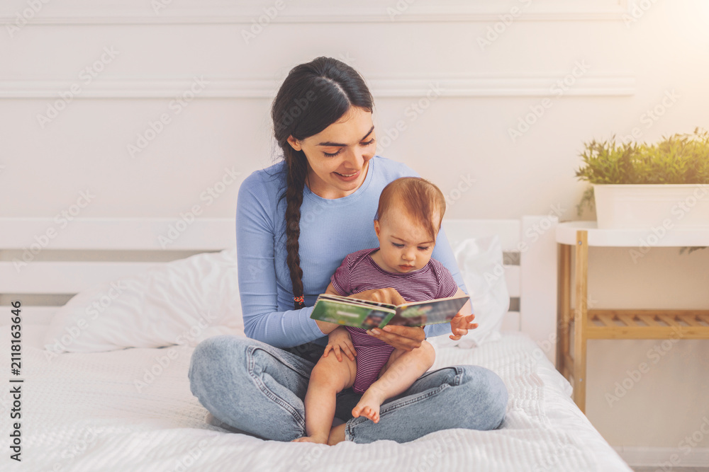 Obraz premium Smiling Hispanic Woman Reading Book To Baby While Sitting On Bed At Home, Enjoying Quality Time Together In Cozy Indoor Setting, Emphasizing Family Bonding And Early Education Concept.