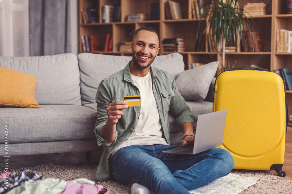 Fototapeta premium Smiling Young Man Sitting On Floor With Laptop And Credit Card In Hand, Happy About Shopping Experience At Home, Surrounded By Luggage And Clothes, Engaged In Online Purchase Process.