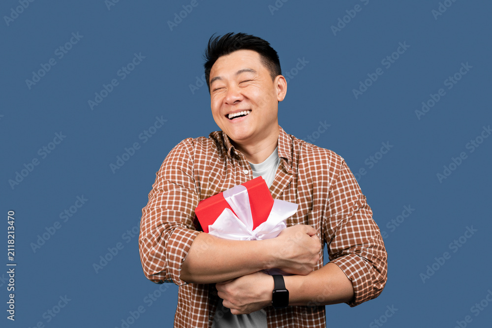 Obraz premium Happy Asian Man Smiling While Holding A Gift Box With Ribbon, Celebrating An Occasion With Joyful Gesture, Blue Background, Indoor Setting, Festive Mood, Celebration Concept.