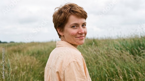 Short-haired redhead young woman with visible facial acne looks back with a knowing smile in windswept grassland, reflecting fearless skin acceptance, open-air freedom, and grounded self-worth