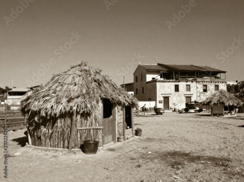 Vintage sepia-toned landscape of a rustic traditional thatched-roof hut in a dusty village courtyard with a historic stone building in the background, antique rural heritage scene.

