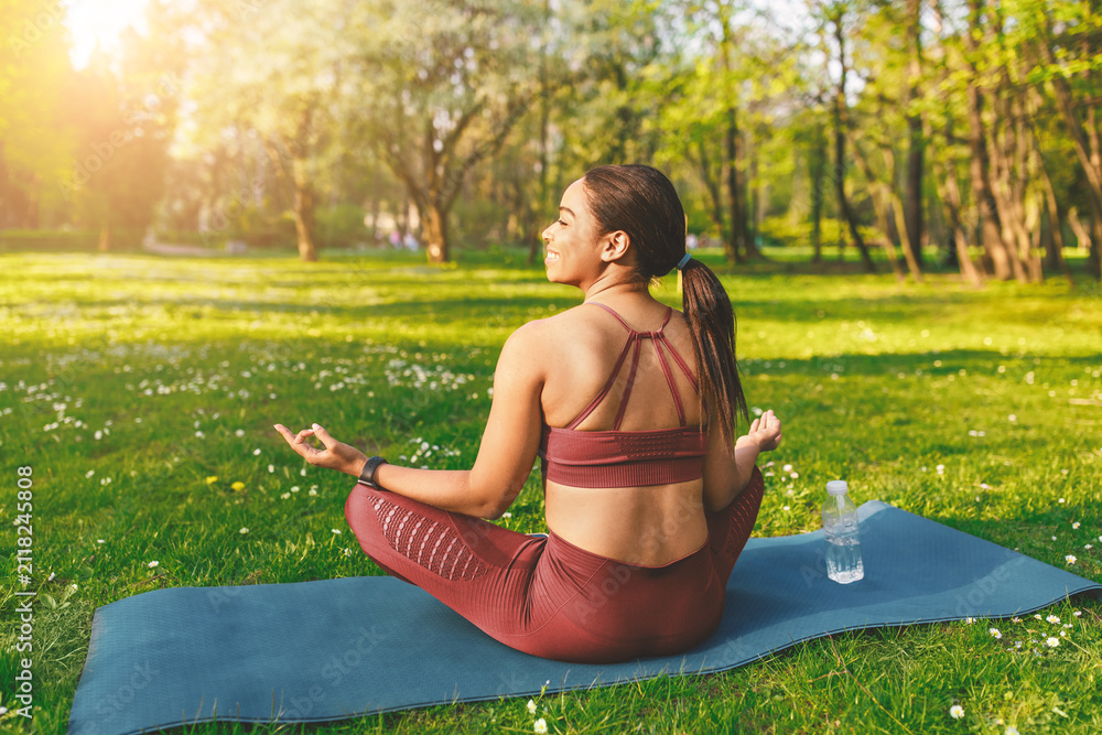 Fototapeta premium Smiling Young Hispanic Woman Practicing Yoga And Meditation In A Park, Sitting On Mat With Calm Expression, Surrounded By Nature. Outdoor Relaxation And Mindfulness Concept.