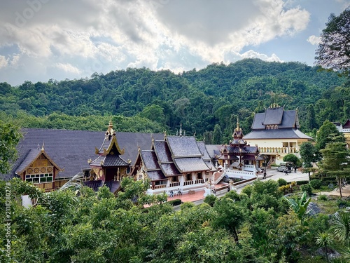 Wide landscape view of traditional Lanna-style temple roofs nestled amidst a vast, lush green mountain forest under a dramatic cloudy sky at Wat Tham Chetawan in Nan province, Thailand.