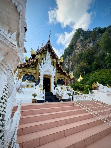 Detailed facade and entrance of a traditional Lanna-style temple building with intricate white wood carvings and a dramatic mountain cliff background at Wat Tham Chetawan in Nan province, Thailand.