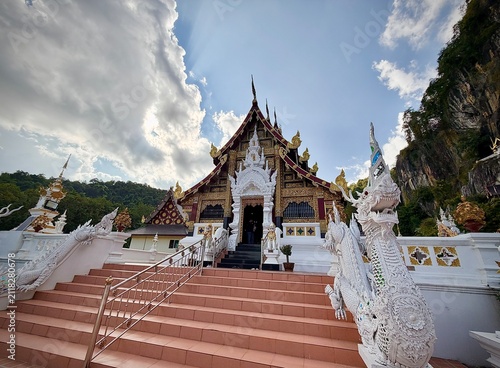 Detailed facade and entrance of a traditional Lanna-style temple building with intricate white wood carvings and a dramatic mountain cliff background at Wat Tham Chetawan in Nan province, Thailand.