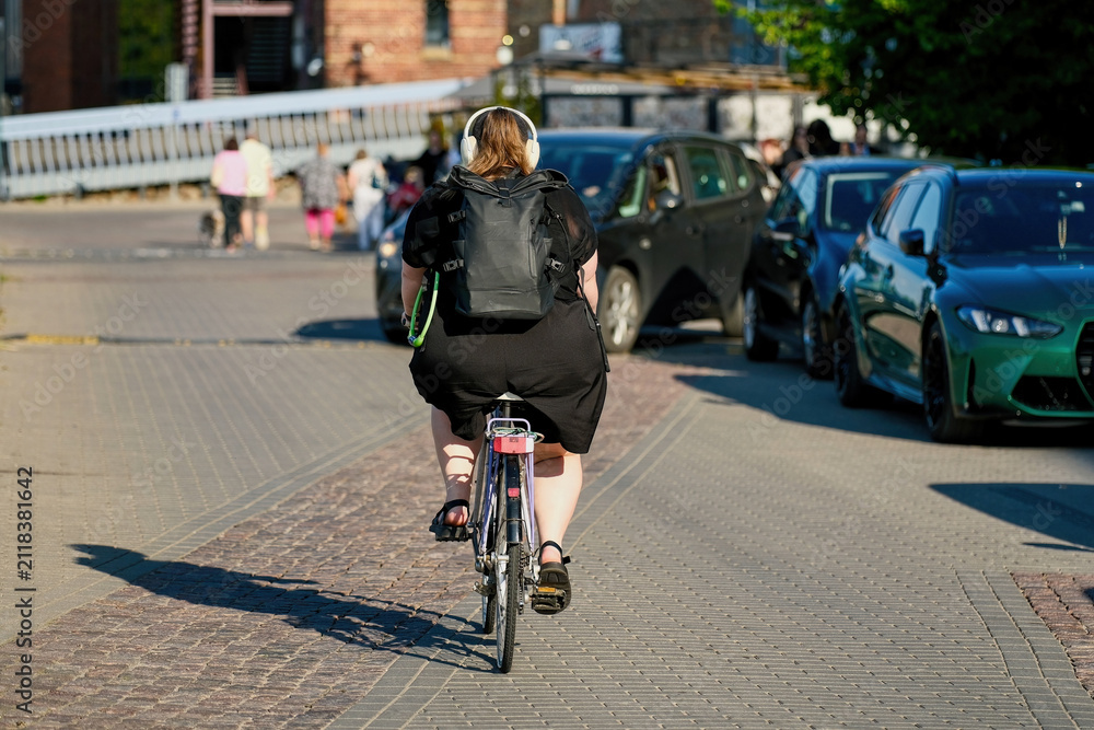 Fototapeta premium Rear view of an overweight woman in headphones riding a bicycle on a city street pavement. Plus size female cyclist commuting on a bike.