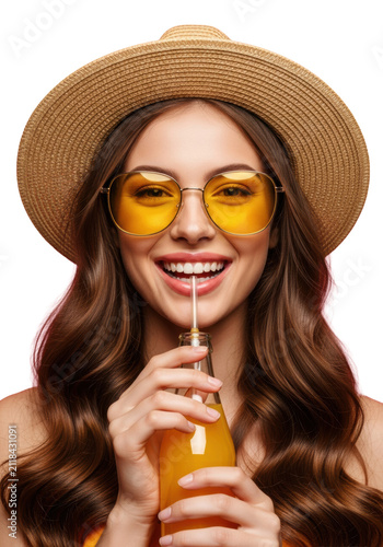 cheerful young woman (20s) in sun hat and reflective sunglasses, sipping orange beverage through a straw from a glass bottle on a vibrant hot pink studio background, clean close-up. concept for