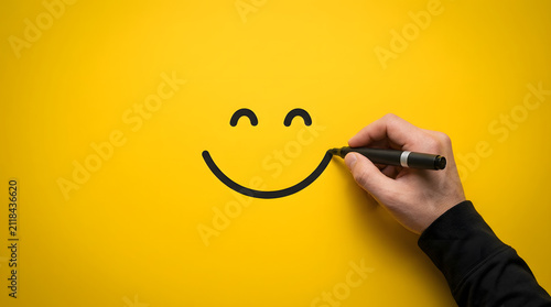 A hand in a black sleeve draws a simple smiley face with a black marker on a vibrant solid yellow background, completing the curved smile line featuring drawing, happy, emotion, bright and colorful