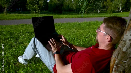 Busy man working on a laptop while sitting on the grass in a city park sitting under tree.