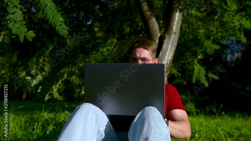 Busy man working on a laptop while sitting on the grass in a city park sitting under tree.