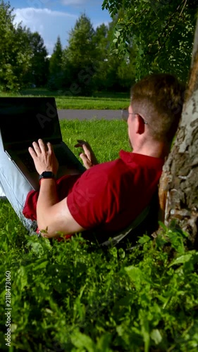 Busy man working on a laptop while sitting on the grass in a city park sitting under tree.