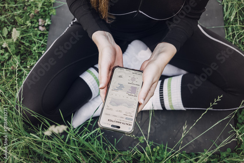 Person sits cross-legged on fitness mat on grass holding smartphone displaying health analytics application. Weekly health summary, wellness data trends, health analytics dashboard, Health report app,