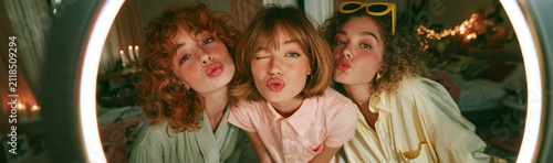Friendship's Fun Moment: Three young women share a playful moment, posing for a selfie with puckered lips and winking eyes, surrounded by fairy lights and a ring light.