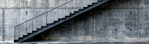 Modern Staircase Against Concrete Wall: A stark metal staircase ascends against a textured concrete wall, reflecting on a wet, polished floor, embodying minimalist design and urban industrialism.