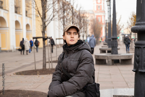 Young man in jacket and cap sitting on city street. Teenager feeling thoughtful in urban environment during autumn day. Lifestyle portrait of student waiting in outdoors public space.