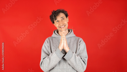 Obraz na plátně Young man clasping hands in pleading gesture against bright red studio background, smiling kindly, expressing hope, prayer, gratitude, or asking for favor in casual grey hoodie