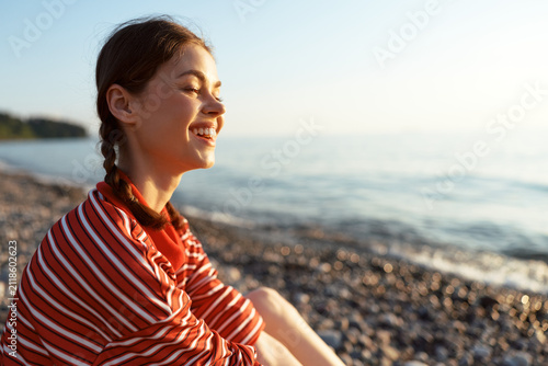 Young woman in a red striped shirt relaxes on a rocky beach, smiling warmly as the sun sets over calm water, her braided hair adds a casual vibe today.
