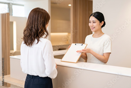 美容クリニックの受付カウンターで問診票を使いお客様に案内や説明を行う笑顔の女性スタッフと接客サービスのイメージ