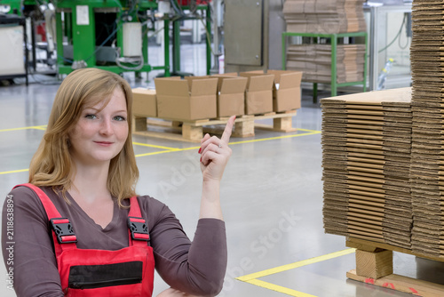 Young female employee in work clothes points to the interior of the assembly hall, which features a designated communication zone and cardboard stored on wooden pallets.
