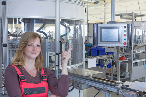 Young female employee in work clothes is pointing to the automatic production line with a control panel.