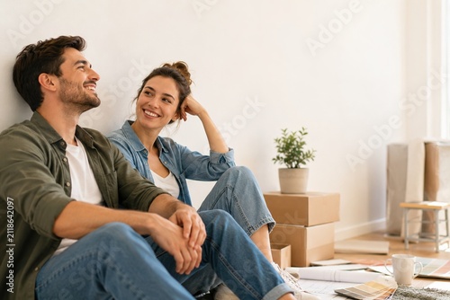 Smiling couple sitting among moving boxes planning home renovation in bright apartment with copy space