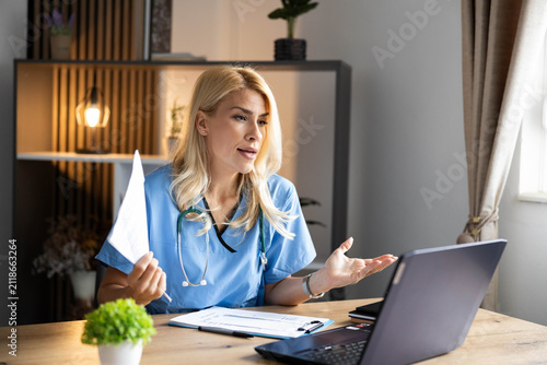 Female doctor in scrubs and stethoscope providing online medical consultation via laptop video call, explaining patient care