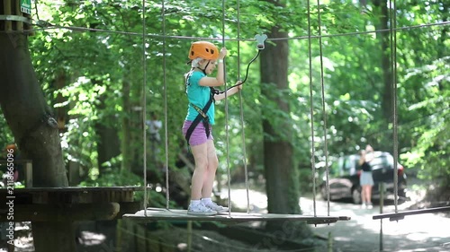 Young girl crossing an obstacle on a ropes course in an adventure park, wearing a helmet and safety harness among trees