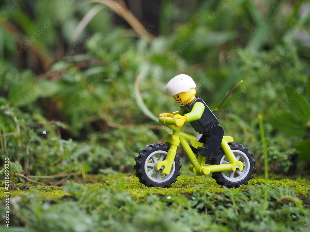 Fototapeta premium Indonesia - May 14, 2022 : Mini toy of action figure at grassland with blurred background. Miniature photography concept. Bicycle at outdoor with green grass conceptual design.