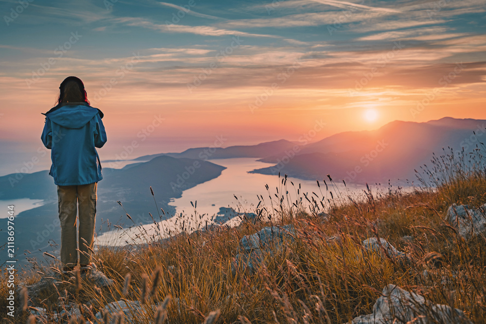 Fototapeta premium Woman standing on mountain top at sunset, looking across scenic bay and mountains, finding freedom and peace in nature