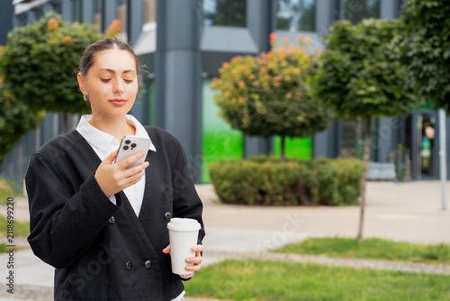 A young businesswoman reading a message on her smartphone while holding a white coffee cup outdoors against a modern building background, representing a corporate lifestyle concept.