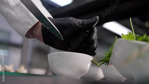 Close up of chef hands in black gloves slicing mushrooms with knife into white bowl. Action moves from slicing to finishing. Professional kitchen setting with green herbs nearby