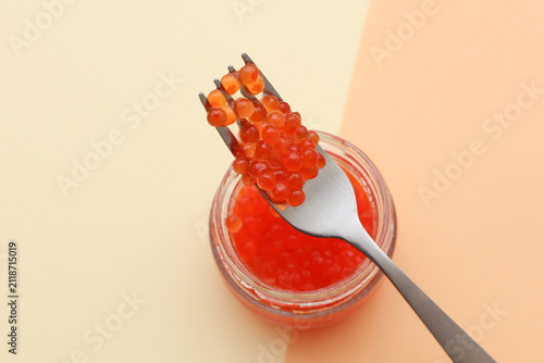 Glass jar of red caviar with fork on a beige background
