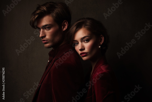 Elegant man and woman posing back to back in burgundy tailored outfits, leaning on each other. Dark and moody fashion shoot