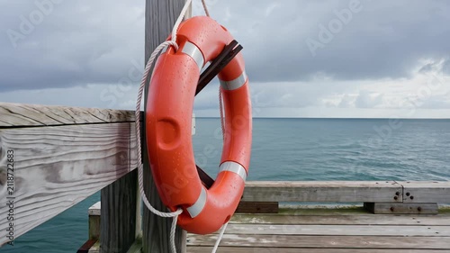 Orange Wet Lifebuoy Hanging on Weathered Wooden Pier Beside with Ocean in the Background