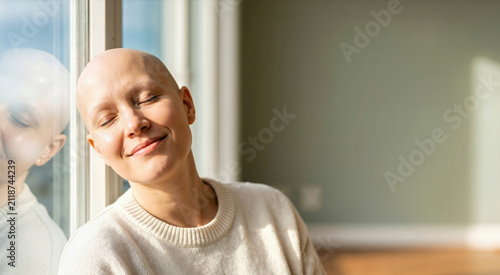Portrait of a smiling bald woman with closed eyes leaning against a window. Happy female alopecia or cancer patient in a sunlit room wearing a cream sweater. Hope and recovery concept
