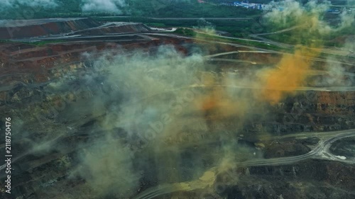 Aerial tracking shot of open pit mine with dust and orange smoke after detonation plumes over terraced excavation roads, mining industry concept.