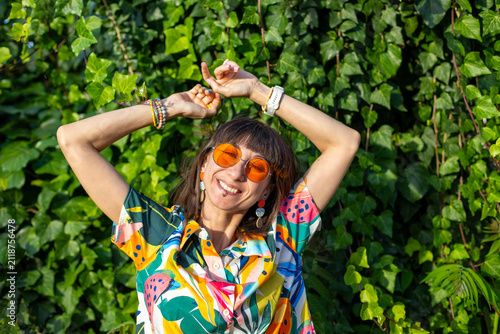 Photography A young woman wearing sunglasses and fashionable accessories stands among the green foliage of the trees